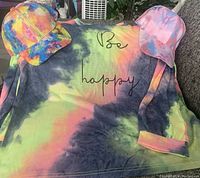 Long sleeve tie dye shirt with 'Be happy' text laid out flat with two tie dye adjustable caps positioned on top on either side of shirt.