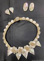 Photo showing the mother of pearl choker necklace with flower and leaf designs alongside two pairs of clip-on earrings on a dark surface.