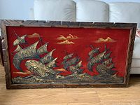 Full view of wooden framed painting showing three Spanish galleons on red felt canvas with gold and black paint and yellow clouds, on hardwood floor with couch backdrop.