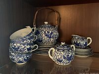Blue and white porcelain Asian style tea set items displayed on shelf showing stacked plates, teapot with wood handle, sugar container and milk container with saucers and cups.