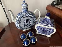 Photo showing round flat blue and white teapot with floral and geometric blue designs, square cookie jar with lid, and four small blue teacups with saucers, on table.