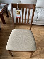 Front view of brown hardwood framed chair with light beige upholstered padded seat, Stakmore tag attached to backrest, placed on wooden floor.