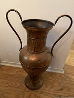 Full front view of a tall copper decorative floor vase with two curved handles, showing the vase's shape and patina finish.