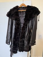 Front view of dark espresso brown leather coat with plush rabbit fur trim along collar and front edges hanging on wooden hanger.