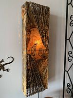 Front view of rectangular decorative wall sconce showing painted hide design and woven stick elements, lit.