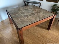 Full view of square dining table with tiled top and solid wood frame with straight legs on wood flooring.