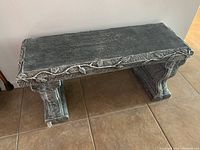 Full view of ornate dark grey concrete or stone bench showing the entire bench with leaf and vine edge detail, curved legs, and textured surface.