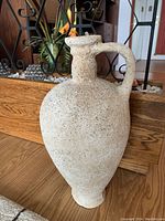Front and side view of the sand-colored textured floor vase showing the handle and overall shape, placed on hardwood floor near decor elements.