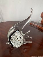 Angelfish glass sculpture viewed from side angle on dark wooden table, showing clear body with speckled black, white, and gold section in center and extended fins and tail.