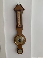 Full view of wooden wall-mounted combination instrument with peaked top. Shows thermometer vertically along the upper section, barometer in the middle circular section, and hygrometer at the bottom.