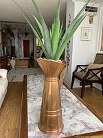 Large copper floor vase with wide top and narrow center, filled with an artificial succulent surrounded by decorative white and black stones, situated indoors on a wood floor.