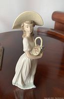 Front and side view of the Nao Spanish figurine showing the young woman in a large wide-brimmed hat holding a flower basket.