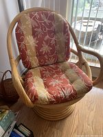 Front view of a light golden brown rattan swivel chair with patterned cushions in rust red and beige tones placed on hardwood flooring near window blinds.