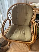 Front angle view showing rattan chair with beige seat and back cushions with button tufting, placed on wooden floor near window blinds.