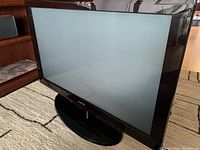 Large Samsung plasma TV in black bezel on oval base stand placed on carpet next to furniture