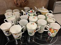 Photo showing a collection of 18 tea and coffee mugs with varied floral and fruit patterns, arranged with two ceramic teapots on a kitchen countertop.