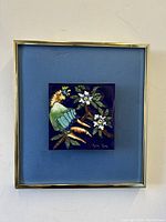 Frontal photo of the framed enamel on metal wall art showing the exotic bird and flowers on blue background, signed by Molly Kates.
