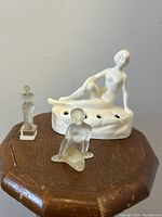 Three nude lady figurines displayed on wooden octagonal surface, white porcelain flower frog reclining and two smaller clear glass nudes in front