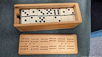Wooden box with the sliding lid removed showing white domino tiles with black pips inside. The cribbage board is shown separately with its rows of holes for scoring.