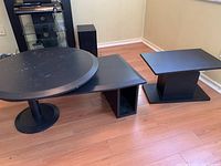 Full view of black round swivel coffee table with an angular base and attached magazine/book rack, next to black rectangular swivel side table. Tables are on wooden floor and show surface scratches.