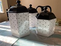 Three hexagonal ceramic cookie jars with black lids and white bodies decorated with black polka dots, three sizes shown together on table with natural light.