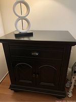 Front view of a black wood end table with single drawer and cupboard below, showing hardware and wood finish.