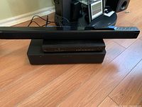 Long black soundbar placed on top of a subwoofer and a DVD player on a wooden floor, showing the full sound system