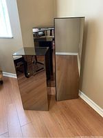 Three mirror stands positioned against beige walls and wood laminate flooring. The stands reflect parts of the room including chair, outlets, and floor details.