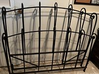 Wrought iron full size headboard standing against a white door, showing vertical bars, curled metal detailing on top, brown-black finish.