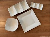 Four white ceramic serving pieces on wooden floor: a bowl, a square platter, a larger triple-sectioned platter, and a smaller triple-sectioned snack tray.