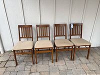 Four wooden foldable chairs with vertical slat backs and beige fabric cushions, aligned side by side on a stone patio.