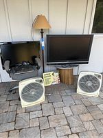 Photo showing two flat-screen TVs, a grey office chair with Dell laptop and cables, a beige floor lamp, two cream box fans, and coffee sign.