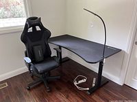 Arozzi gaming chair next to L-shaped black desk with integrated power strip and gooseneck lamp