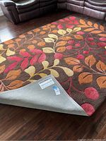 Floor rug laid out on wood floor showing brown base with red, orange, and cream leaf pattern. Corner flipped to show underside and label.