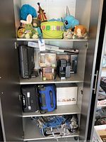 Full view of cabinet shelves with all items in lot including soft toys, coffee machine, camping coolers, hiking backpack, and boxed lights.