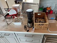 Kitchen countertop displaying a multi-tier spiral stainless steel fruit basket, a Super Chef 2000 manual vegetable slicer with box, an electric coffee grinder, a wicker basket holding spoons, and a large serving tray.