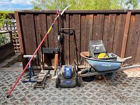 Full lot including lawnmower, wheelbarrow, red pruning tool and floor jack against wooden fence on paved ground.