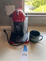 Front and side views of the red and black Nespresso VertuoLine coffee maker with water reservoir and power cord visible. Machine accompanied by green ceramic mug.