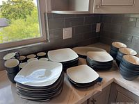 Dinner and salad plates stacked with mugs and bowls on countertop under window.
