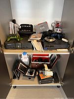 Similar angle showing Xbox and PlayStation 2 consoles with games manual, green wired controller, joystick controller and Wii manual on top shelf.