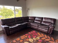 Front view of the large dark brown sectional leather couch positioned by a window, showing the seating area and armrest, along with a colorful rug beneath.