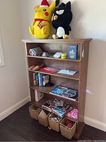 Photo displaying wooden bookshelf with two plush toys on top, various books and magazines on shelves, and woven baskets placed at bottom.