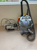 Photo showing gray Shop Vac with hose, handle, and vacuum pump on floor near wall