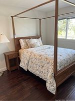 Twin wooden canopy bed frame with bedding, wooden nightstand, and white table lamp near window.
