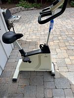 Photo of upright exercise bike positioned outside on stone patio; shows seat, pedals, handles, and display console.