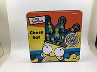 Front view of the Simpsons tin box chess set showing a window with some of the character pieces inside and colorful Simpsons artwork on the lid.