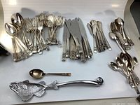 Full overview of the cutlery set laid out on a white surface showing spoons, forks, knives, butter knives and serving spoons including a fish-shaped serving spoon.
