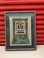 Framed vintage textile art with meditative figure in center mount and decorative fabric border, leaning against a red background.