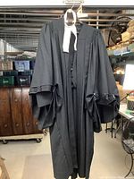 Full view of the black vintage barrister robe hanging, showing pleated and buttoned wide sleeves and white neckband.