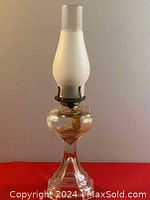 Full view of the antique oil lamp standing upright on a red surface. Shows the clear glass base, brass burner, and frosted glass chimney.